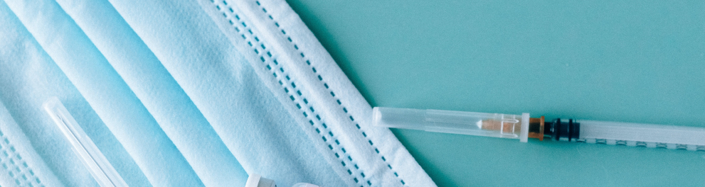 Imagem de seringa, agulha de injeção, frasco de vidro com rótulo escrito covid-19 e uma máscara de prevenção à doença. Todos os itens estão em cima de uma superfície azul
