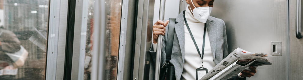 Jovem mulher negra está usando máscara de prevenção à covid, em pé no interior de um vagão de metrô e lendo um jornal