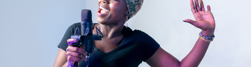 Uma mulher negra com turbante na cabeça está cantando segurando um microfone com a mão direita