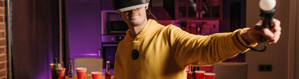 Um jovem branco vestindo um moletom amarelo está dentro de uma sala com um microfone em cada mão e um óculos de realidade virtual no rosto