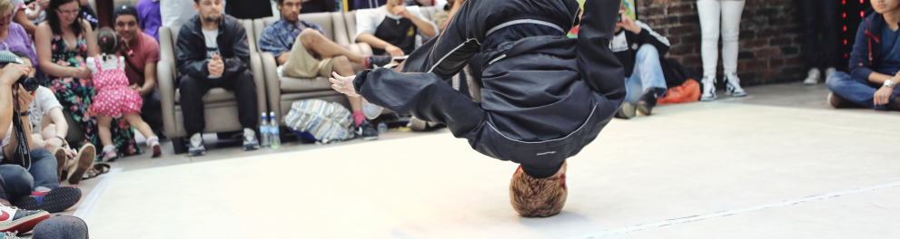 Várias pessoas ao redor da imagem estão olhando para um espaço aberto no centro, onde há um rapaz fazendo um passo de break dance, com a cabeça no solo e as pernas para cima