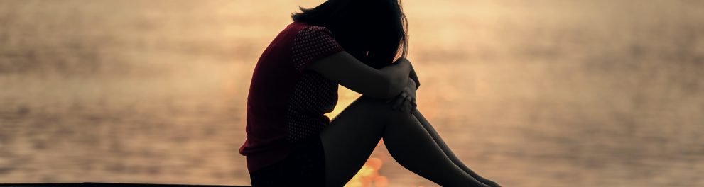 Imagem de uma menina de cabelos pretos e lisos triste sentada no que parece ser um deck de madeira. Ela está triste, com os braços apoiados nos joelhos e a cabeça baixa encostando nos braços. Ao fundo tem uma lagoa ou mar.