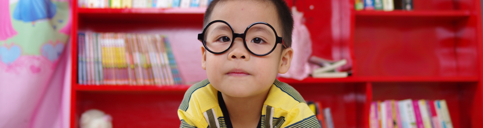 Um menino oriental de cerca de 4 anos de idade e óculos de grau de armação preta e redonda está olhando para quem  bateu a foto. Ele está apoiado em uma mesa branca, com um livro à sua frente