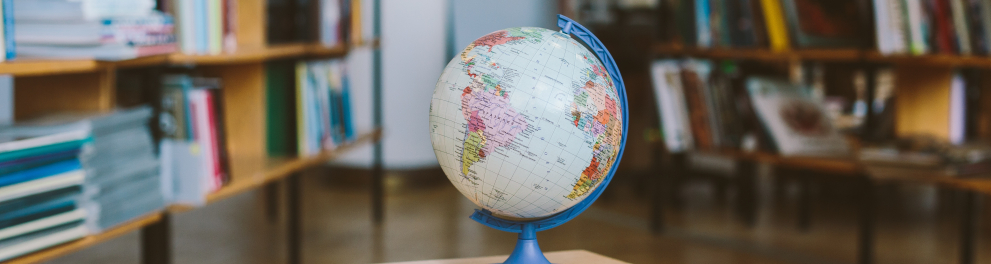 Um globo em miniatura está posicionado em cima de uma mesa de madeira e, ao fundo, podemos ver que o ambiente é uma biblioteca