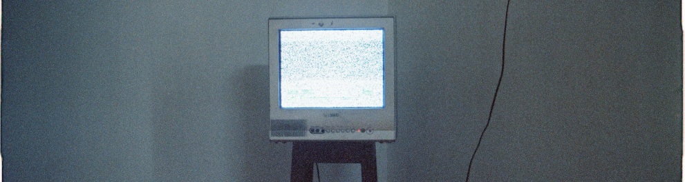 Imagem de uma televisão ligada e colocada em cima de um banco no canto de um quarto.