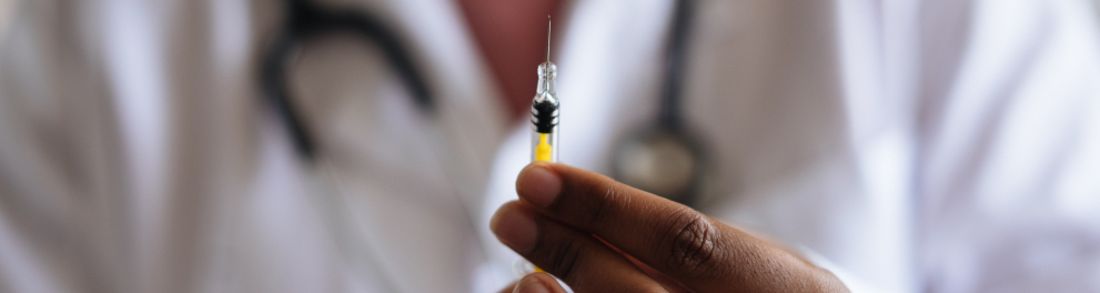 Imagem das mãos de um homem negro segurando uma injeção. Ao fundo, embaçado, podemos notar que trata-se de um médico, pois podemos ver o jaleco e o estetoscópio pendurado em volta do pescoço
