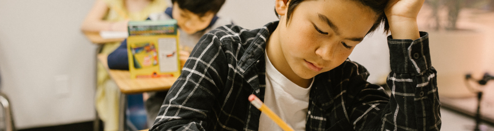 Garoto oriental em sala de aula com outras duas crianças sentadas atrás dele, na fileira. O menino da frente está escrevendo algo no caderno com a mão direita e a mão esquerda está na cabeça