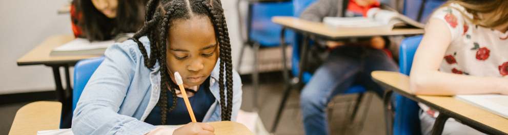 Três crianças estão sentadas nas cadeiras de sala de aula anotando alguma coisa em seus respectivos cadernos. O foco central da imagem é em uma menina negra de tranças