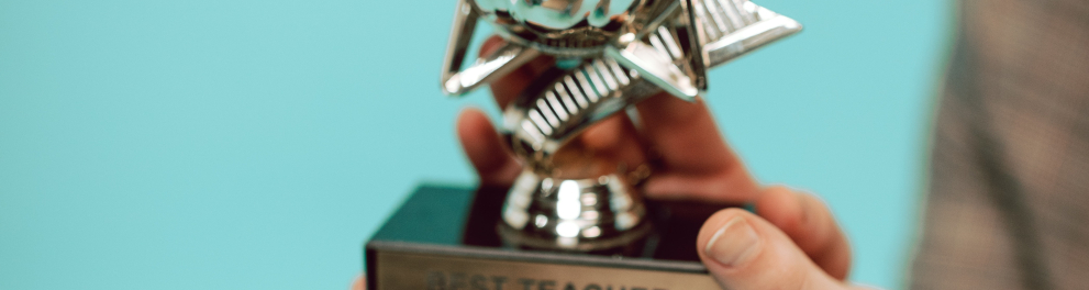 Close em um troféu sendo seguro por duas mãos brancas e com a grafia de "best teacher" na parte inferior do objeto