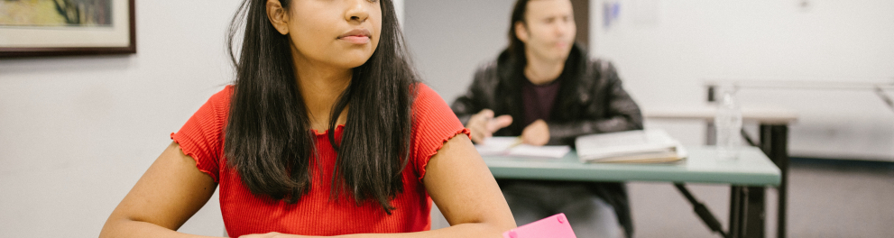 Uma menina adolescente está sentada em sala de aula com os braços sobre a mesa, em cima de material escolar. Ela parece estar prestando atenção em algo à sua frente, que não podemos ver, mas deduzimos ser o professor lecionando