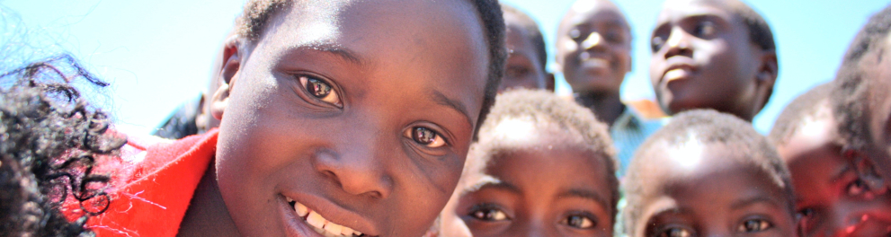 Imagem de crianças negras, possivelmente africanas, olhando de pertinho para a câmera que as fotografou. Elas demonstram curiosidade e estão sorrindo meio sem entender do que se trata