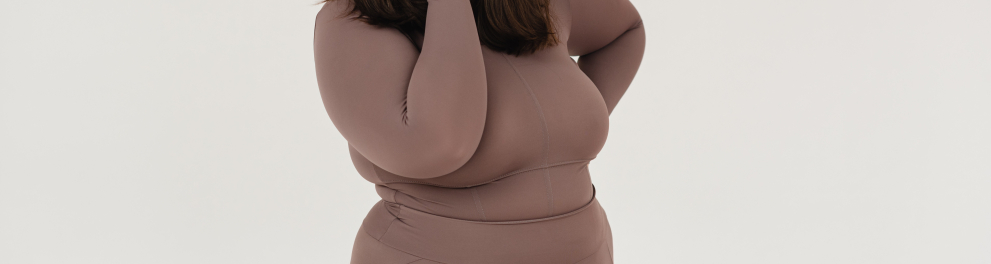 Uma mulher acima do peso está sorridente com a mão direita no rosto e a mão esquerda para trás do corpo em um fundo branco. A mulher está com calça e uma blusa de cor meio cinza