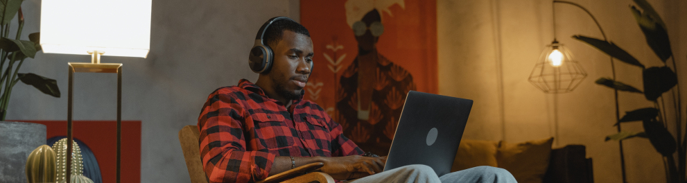 Um homem negro de calça jeans e camisa social xadrez está sentado em uma cadeira em sua sala de estar com um notebook no colo e um headphone na cabeça