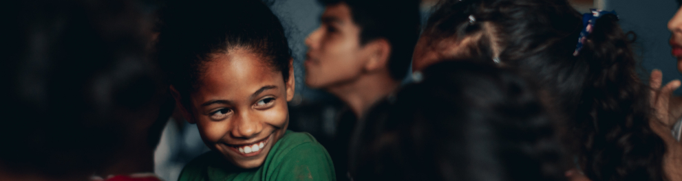 Imagem closada em uma menina negra sorrindo em um local que parece ser uma sala de aula. Podemos ver as cabeças de outros estudantes