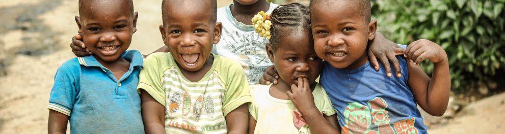 Cinco crianças negras estão lado a lado abraçadas e posando sorridentes para quem bateu a foto. Elas aparentam ser bem humildes