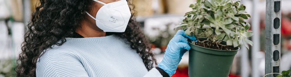Mulher negra de cabelo cacheado está de luvas e máscara de prevenção à covid mexendo um vaso de plantas de uma loja