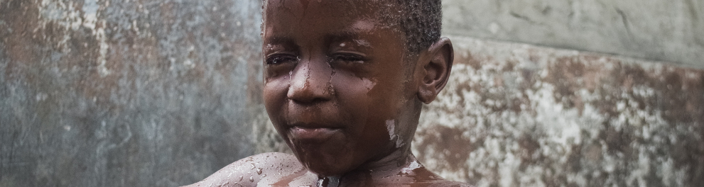 Imagem de um menino negro, de cerca de 4 anos, sentado em uma bacia preta e sendo molhado por água que cai de um pote transparente em sua cabeça. O pote é seguro pelas mãos femininas de uma adulta, provavelmente a mãe do menino