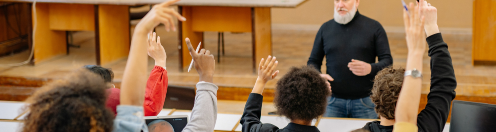 Alunos em uma sala de aula que parece um audiório estão levantando as mãos querendo fazer perguntas ao professor, que está de camisa preta, calça jeans e cabelo e barba grisalhos no centro da imagem