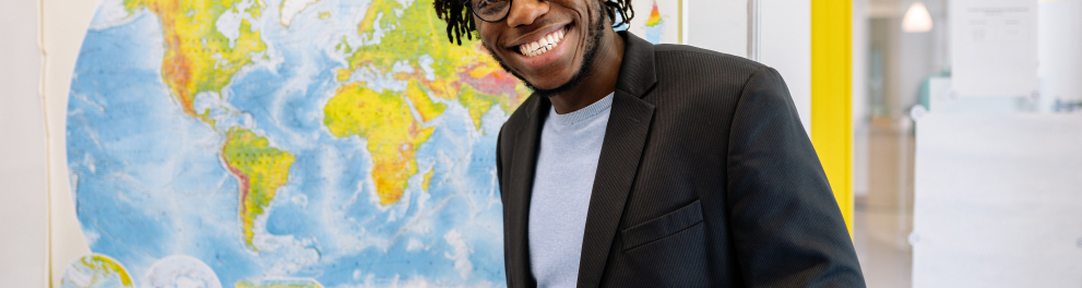 Homem negro de paletó, óculos de grau e cabelo de dread está sorrindo para quem fez a foto e segurando uma caneta com a mão direita e um bloco de papel com a mão esquerda.