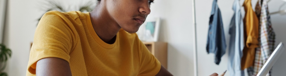 Uma pessoa negra trajando uma camisa amarela e relógio no pulso esquerdo está sentada e mexendo em um tablet branco com a mão direita