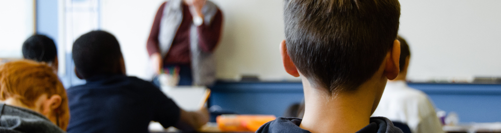 Imagem de alunos crianças de costas para quem bateu a foto. As crianças estão sentadas em suas cadeiras em sala de aula e, ao fundo, dá para vermos o professor em pé em frente ao quadro
