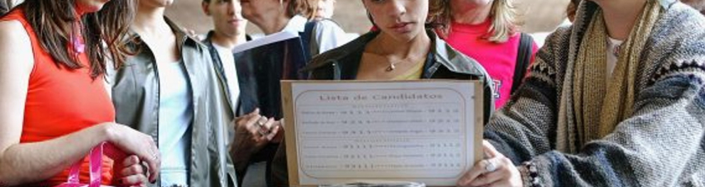 A imagem mostra um grupo de mulheres observando uma lista de candidatos da eleição