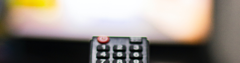Imagem mostra uma mão segurando um controle remoto em frente a uma TV