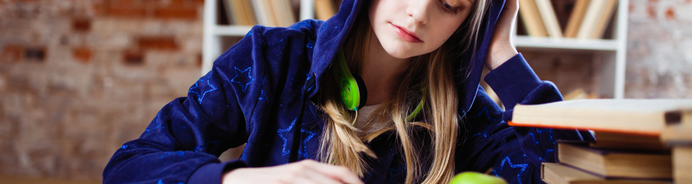 Uma menina loira vestindo um casaco azul com capuz está sentada lendo um livro. Ao lado do livro, há uma pilha de outros livros e uma maçã verde está em cima do livro aberto que ela lê