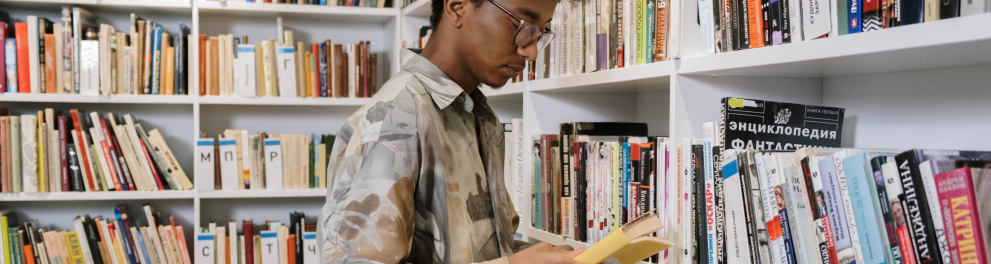 Um adolescente negro, alto, magro e de cabelo escuro arrepiado está em uma biblioteca com um livro em mãos e de perfil para quem bateu a foto. Ao seu redor há várias prateleiras com outros livros