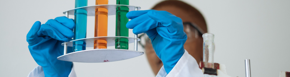 Um cientistas está segurando um recipiente contendo três cilindros de vidro na altura de sua cabeça. Cada cilindro tem uma cor (azul, laranja e verde) e o profissional está olhando atentamente
