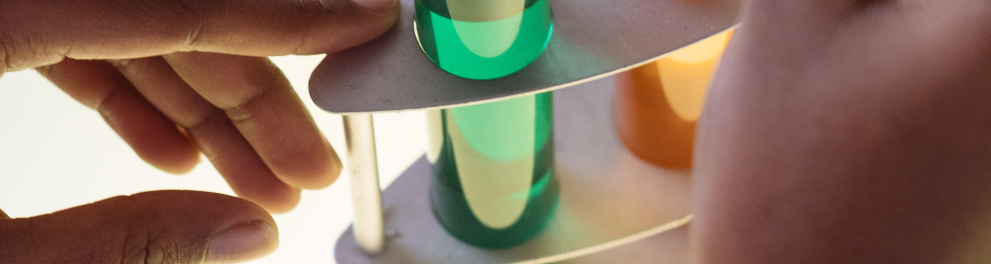 Dois cilindros de vidro contendo líquidos coloridos (verde e laranja) estão em um recipiente e sobre uma mesa branca. Há as mãos de uma pessoa branca retirando parte do líquido laranja com um conta gotas