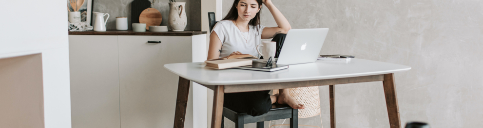 Uma jovem mulher branca, de cabelos lisos, soltos e escuros está sentada em uma cadeira em frente à uma mesa com seu notebook aberto. Ela parece preocupada com algo, inclusive coçando a cabeça com a mão esquerda