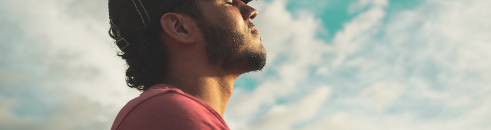 Homem moreno está de perfil para quem bateu a foto. Ele está de olhos fechados e parece respirar fundo ao ar livre