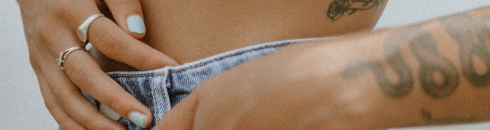 Imagem closada no abdômen de uma mulher branca. Ela está com a mão direita na barriga e a mão esquerda no bolso da calça jeans