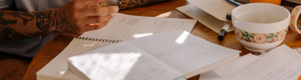 Imagem mostra uma mesa de estudos e uma mão segurando caneta escrevendo em caderno.