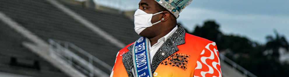 Imagem de um homem negro, obeso, trajando um paletó colorido, coroa na cabeça, máscara de prevenção à covid no rosto e uma faixa azul escrito "rei momo 1° e único 2020"