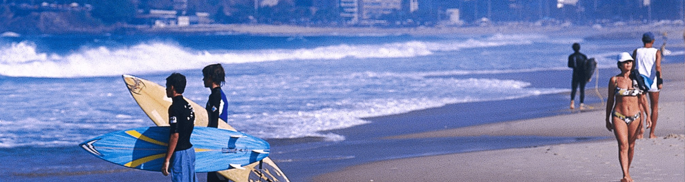 A imagem mostra uma praia, com pessoas passeando na beira do mar e dois surfistas olhando para a água