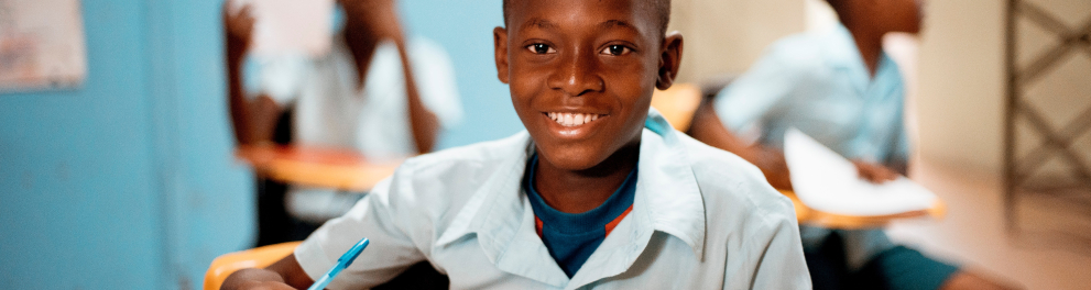 Menino negro esta sentado em sala de aula segurando um caderno com as duas mãos e sorrindo para quem bateu a foto
