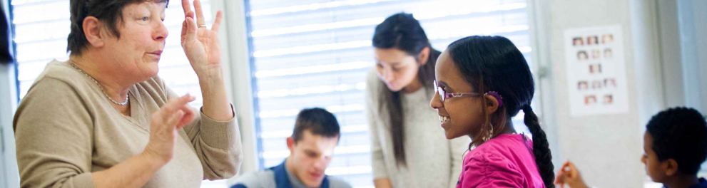 Uma sala de aula com estudantes interagindo e uma aluna conversando com a professora