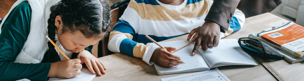 Duas crianças estão diante de livros e cadernos. Uma delas, uma menina, escreve, enquanto o menino, com um lápis na mão, recebe orientação da professora., da qual não vemos a cabeça, apenas braço, mão e parte do tronco.