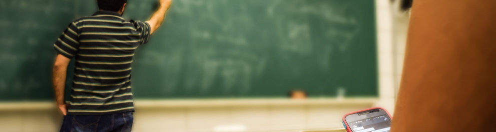 Um homem (possivelmente, um professor) escreve no quadro em uma sala de aula. Em primeiro plano, uma estudante escreve no celular. Dela, só vemos o braço e um pedaço da perna.