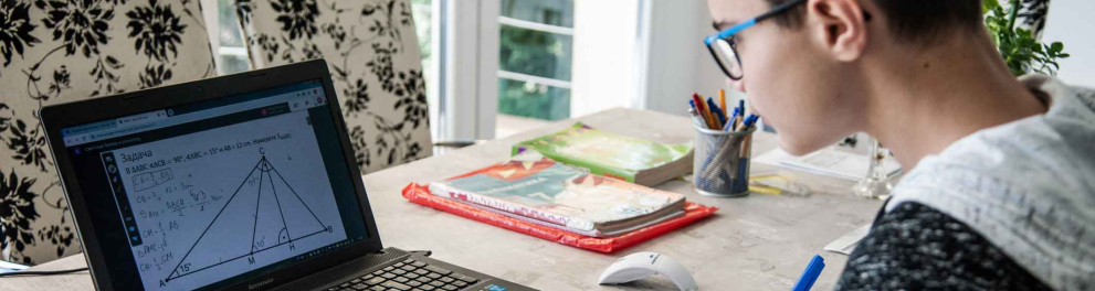 Um jovem, que usa óculos, está escrevendo algo em uma folha branca enquanto observa o que está na tela do laptop.