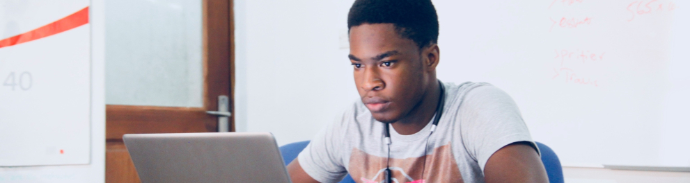 Jovem rapaz negro está sentado e olhando atentamente para o notebook aberto à sua frente.