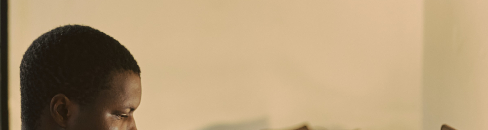 Um jovem rapaz negro está sentado lendo um livro e de perfil para quem bateu a foto. Ele está dentro de um quarto.