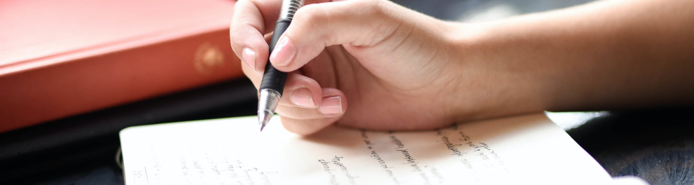 Mão direita de uma pessoa branca está escrevendo algo em um caderno