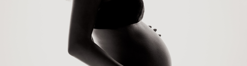 Uma mulher grávida de perfil para quem bateu a foto. Ela está com as duas mãos na barriga e a iluminação do local é fraca, oq eu deixa a foto num estilo preto e branco