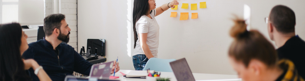 Pessoas sentadas ao redor de uma mesa branca de reunião estão olhando para o quadro branco ao lado, onde há vários post its coloridos colados e uma mulher em pé de cabelos longos e pretos está falando algo enquanto aponta para os post its