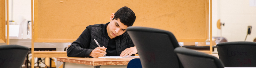Um jovem adolescente de cabelos curtos e escuros está sentado em sala de aula escrevendo alguma coisa em uma folha. Parece estar realizando uma prova