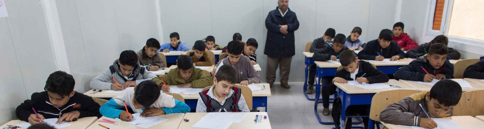 Uma sala de aula com diversos estudantes fazendo uma tarefa enquanto um professor os observa.