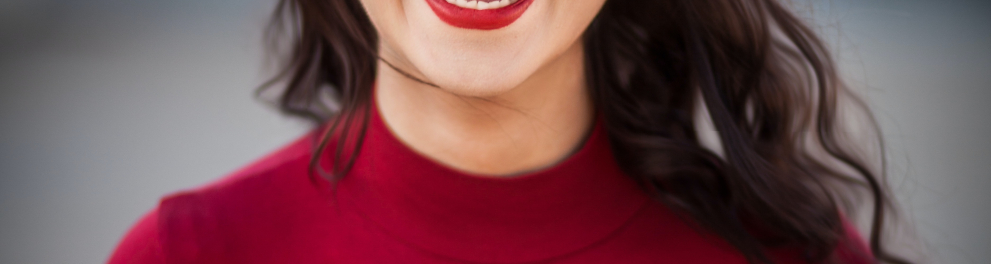 Jovem mulher branca de batom vermelho e cabelos escuros e longos está vestindo uma blusa vermelha de mangas longas e sorrindo com muita felicidade para quem bateu a foto